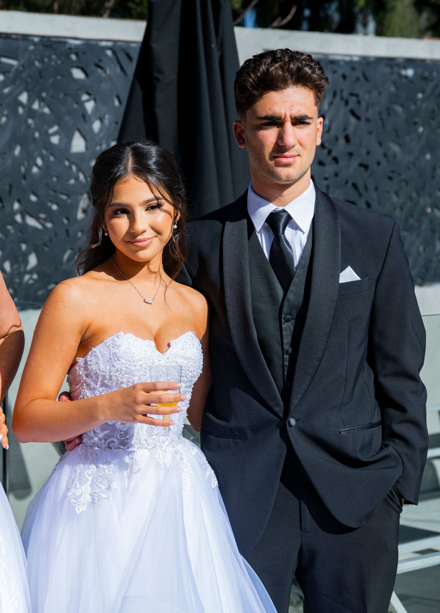 A couple in elegant wedding attire poses together outdoors, with the bride wearing a flowing white gown.
