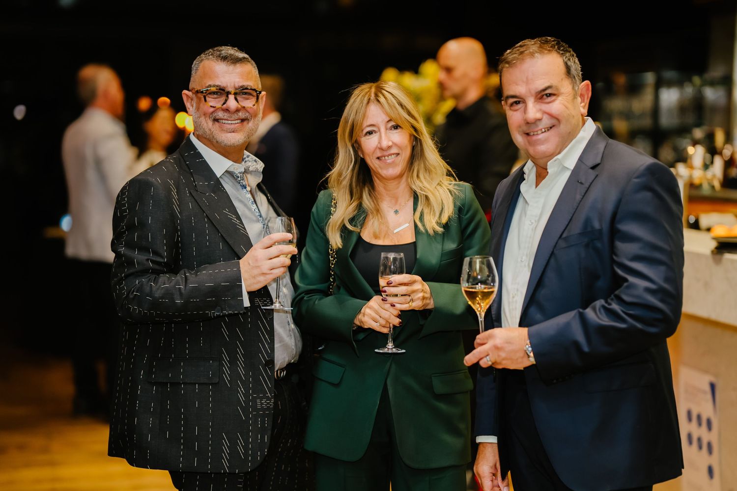 Three people in business attire holding wine glasses at an upscale social event in a dimly lit venue.