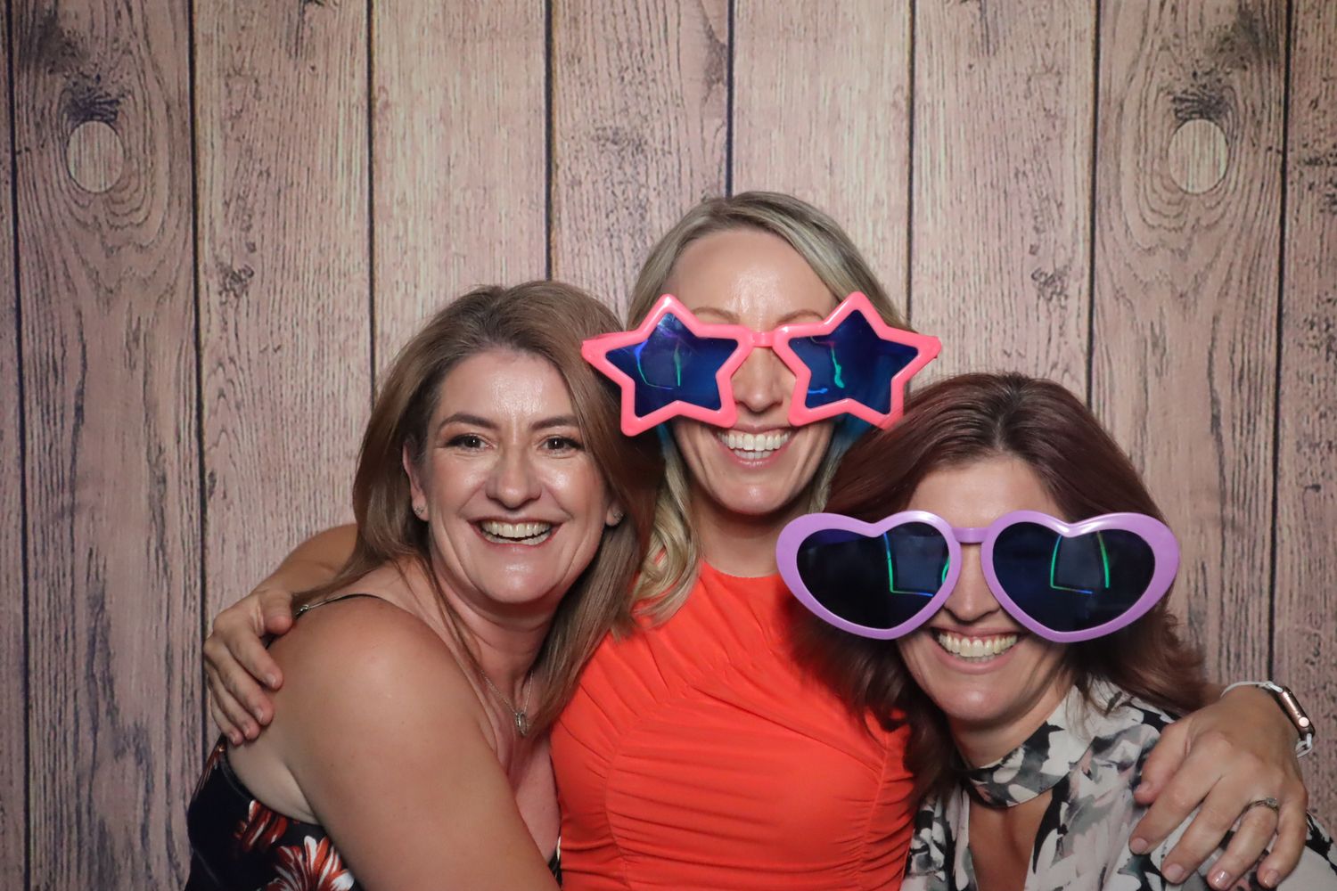 Friends having fun with heart-shaped sunglasses in photo booth against wooden backdrop.