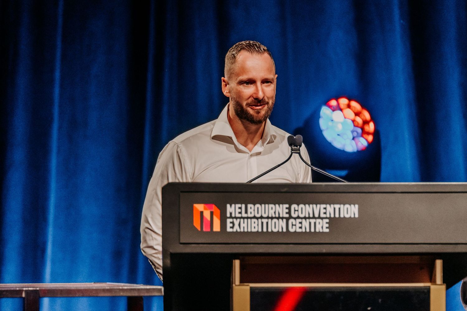 Professional speaker presenting on stage at the Melbourne Convention and Exhibition Centre with blue lighting.