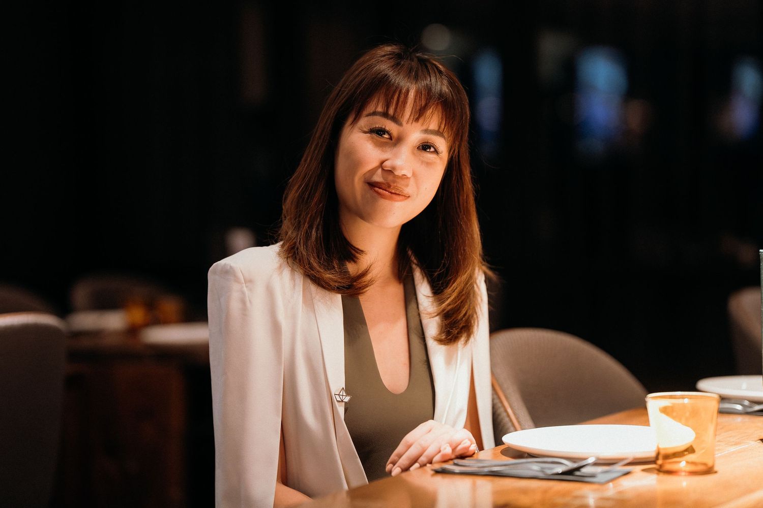 A person in a white blazer sits at a restaurant table with a warm ambient lighting setting.
