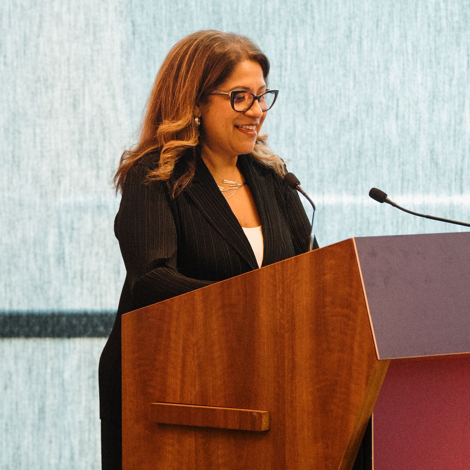 Speaker at podium wearing black blazer and glasses gestures while delivering presentation at professional event.