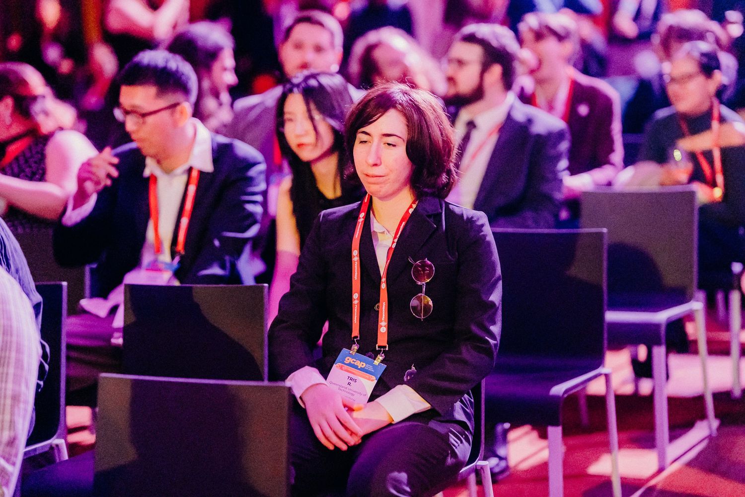 Conference attendees sit in a dimly lit auditorium with pink and purple lighting during a professional event.