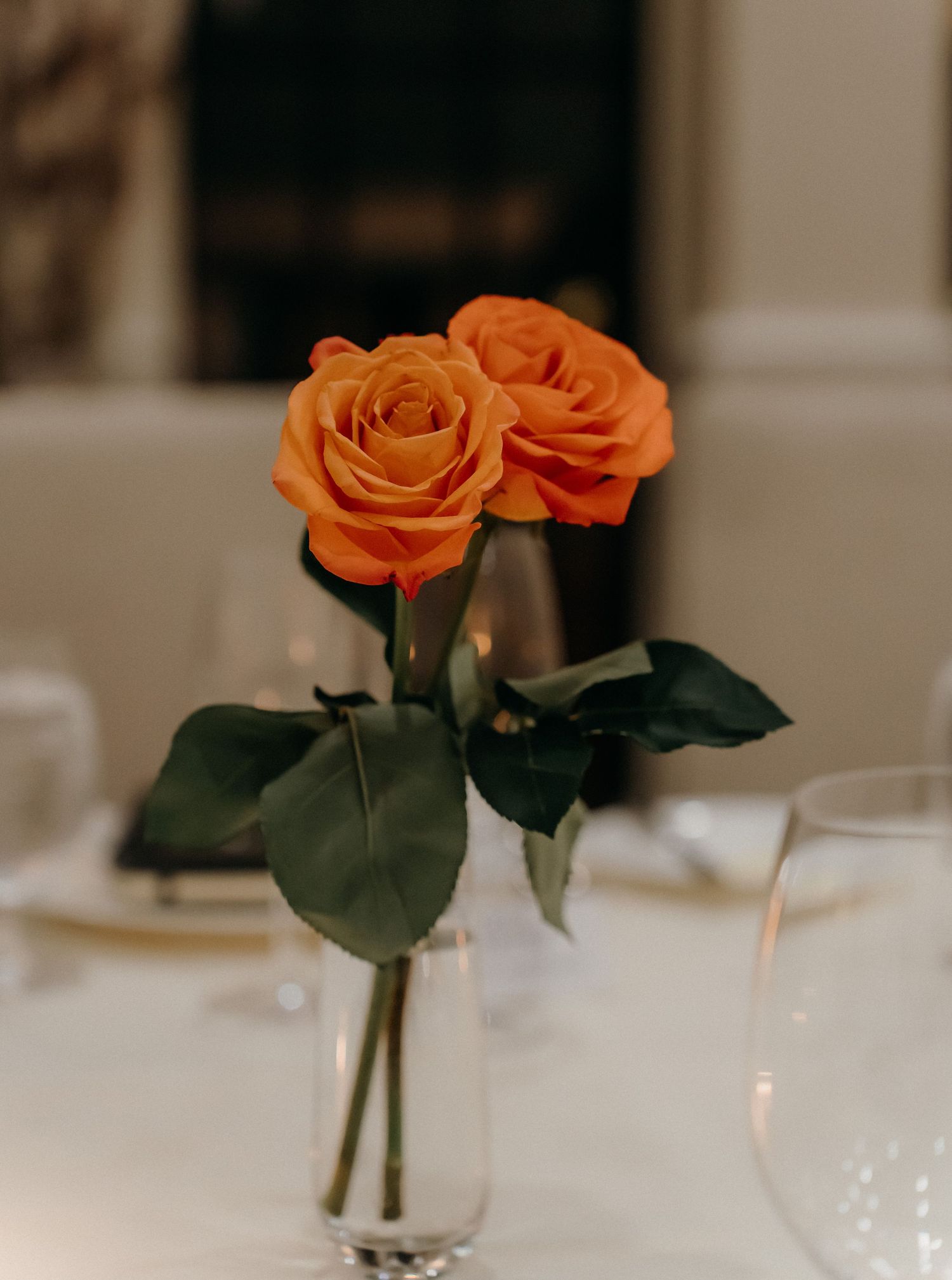 Two orange roses with green leaves arranged in a small glass vase on a white tablecloth for an elegant table setting.