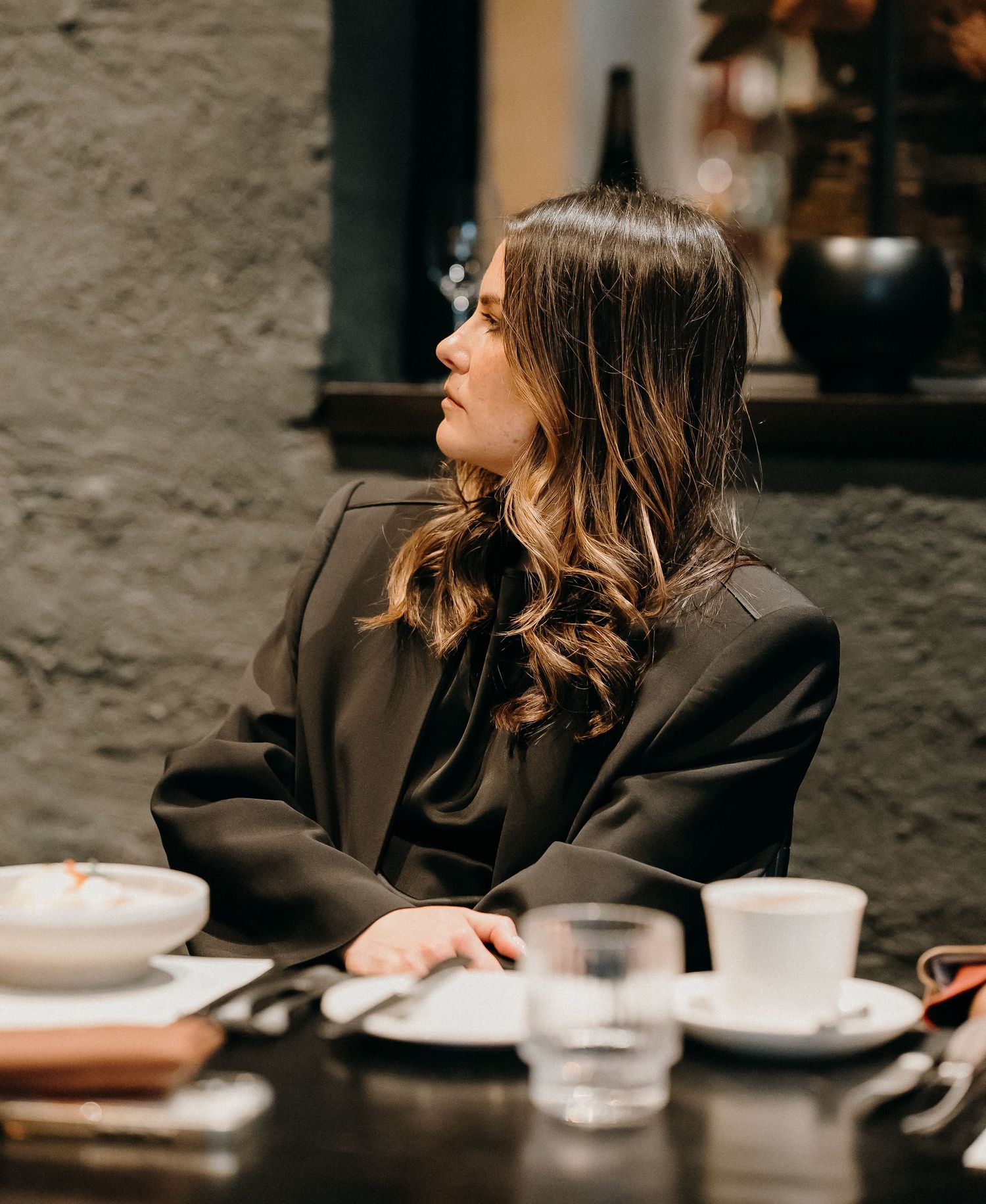 Person in dark blazer sitting at restaurant table with plates and glasses in moody lighting.