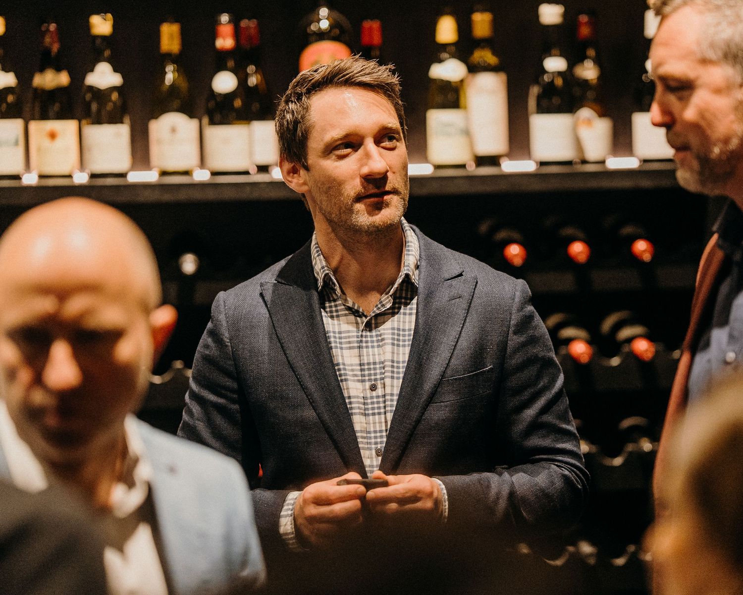 People in business attire gathered around a wine bar with bottles displayed on shelves in the background.