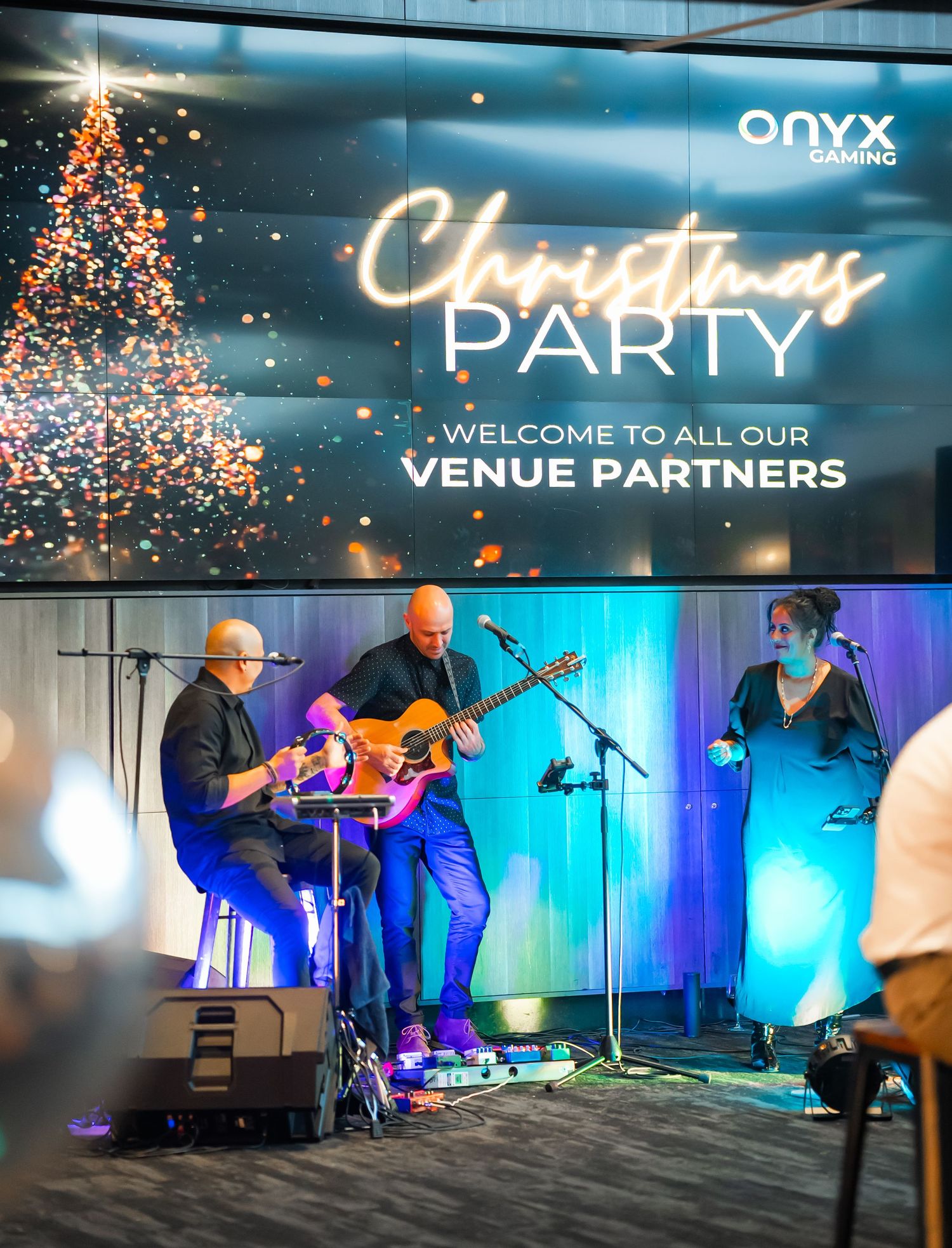 Musicians perform on stage with blue lighting and venue partners branding displayed at a corporate event.