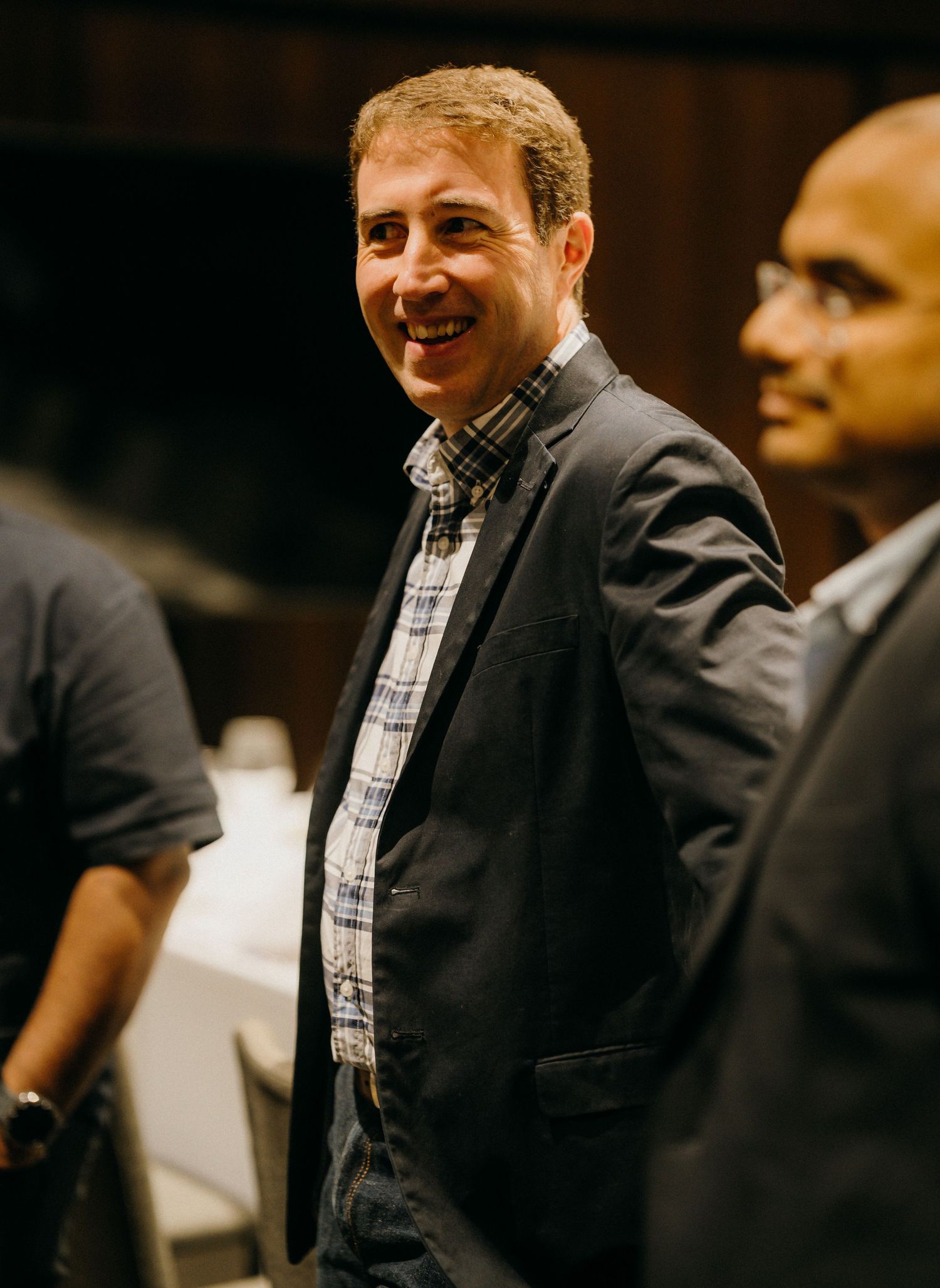 Smiling man in blazer and plaid shirt at an indoor event, with other attendees visible in background.