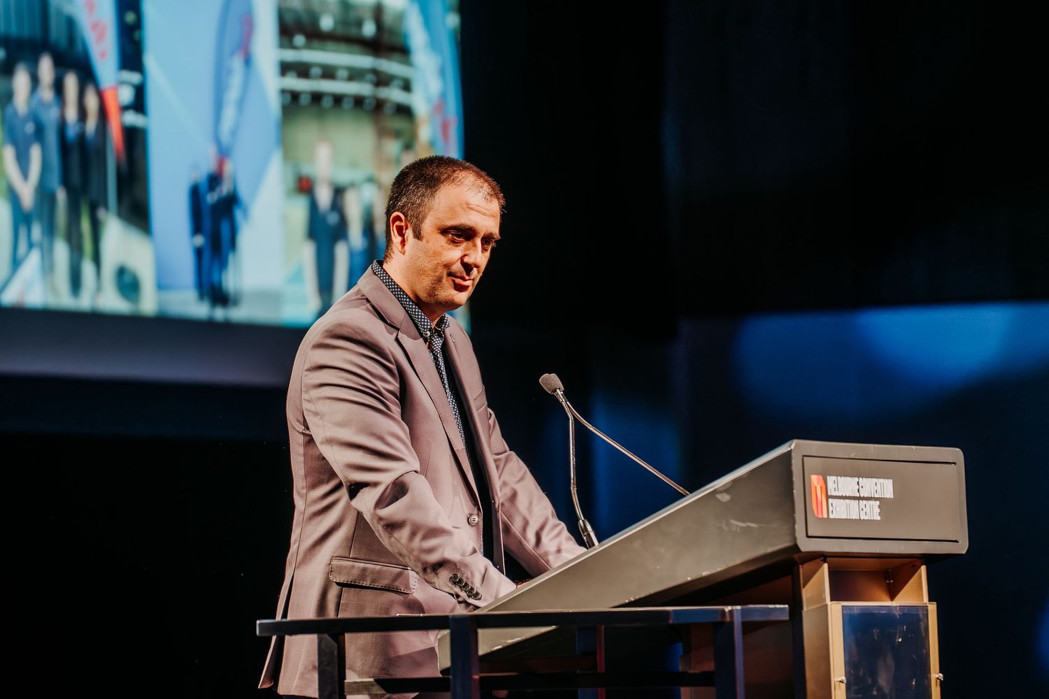 A speaker stands at a podium giving a presentation in a dimly lit auditorium.