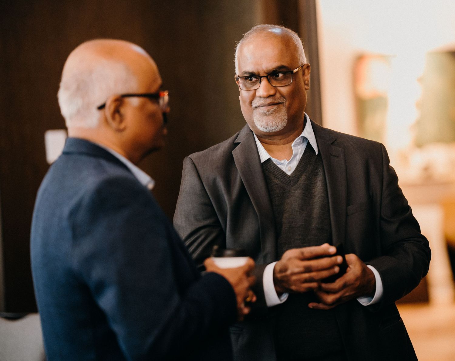 Two businessmen in professional attire having a conversation while one holds a mobile device in an office setting.