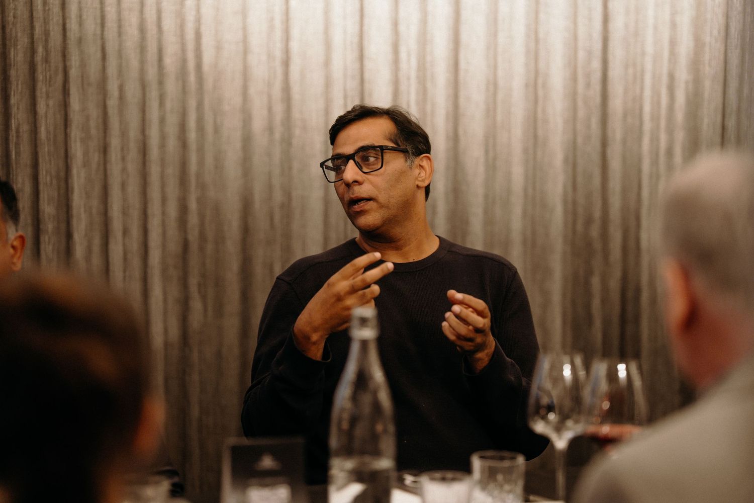 A group of people gather around a table at a restaurant, with wine glasses and dim lighting creating an intimate atmosphere.