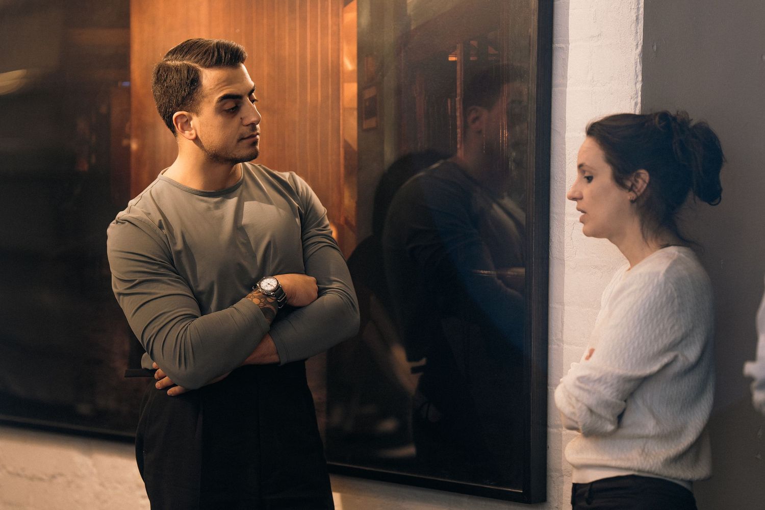Two people having a serious conversation while standing in a hallway with reflective surfaces.