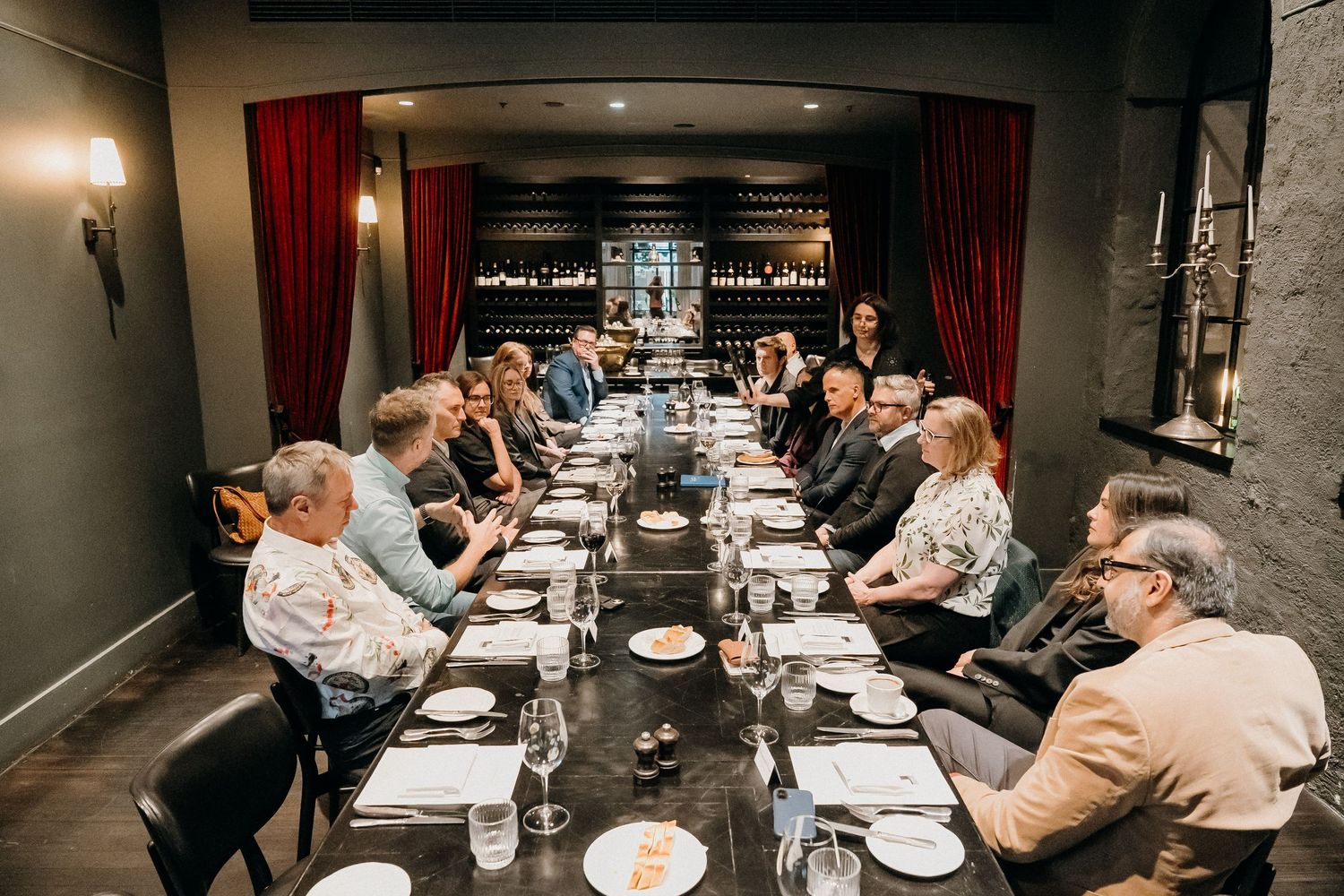 Business professionals seated at a long conference table during a meeting in a dimly lit room with red curtains.