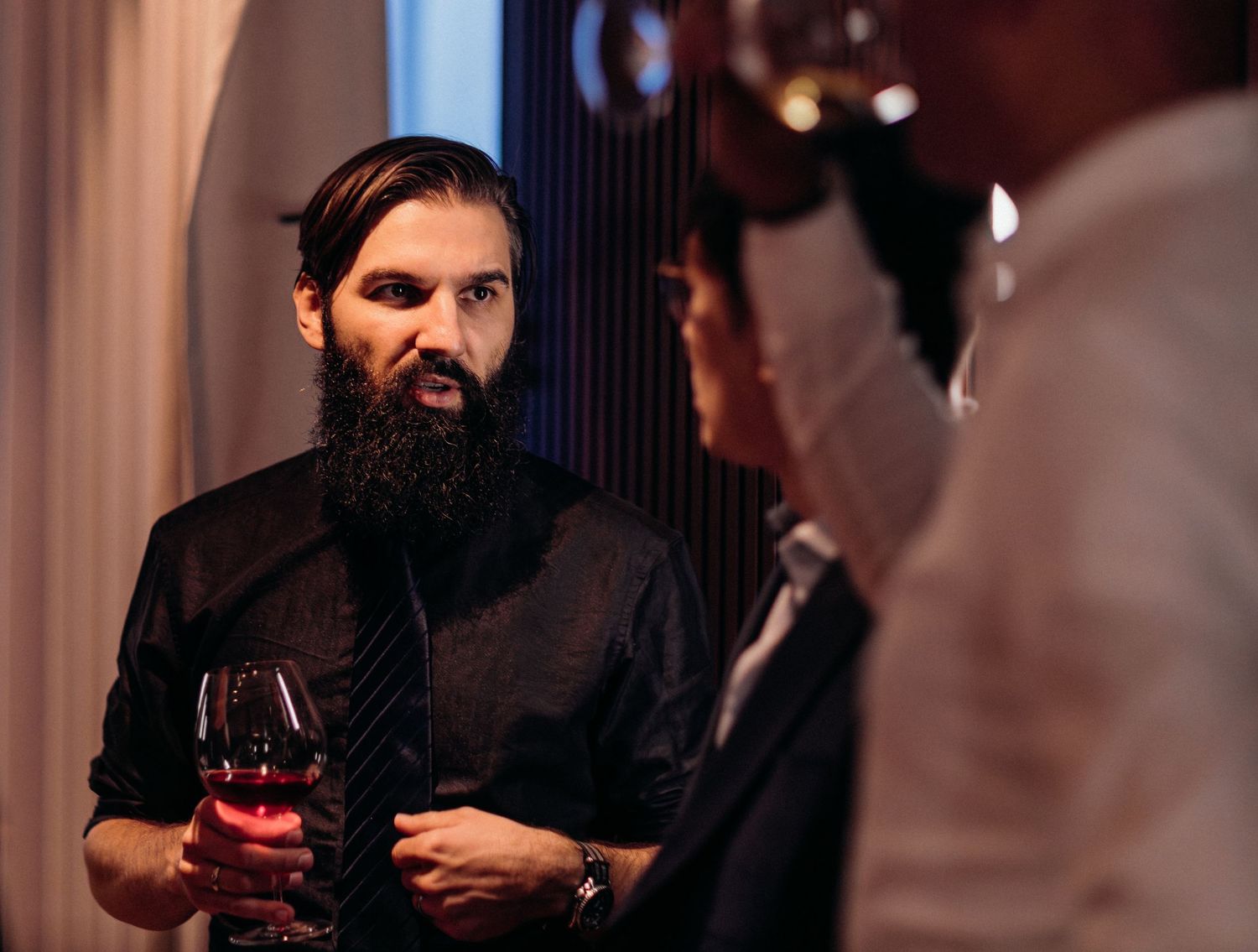A bearded man in dark clothing sits at a dimly lit bar with a glass of red wine.