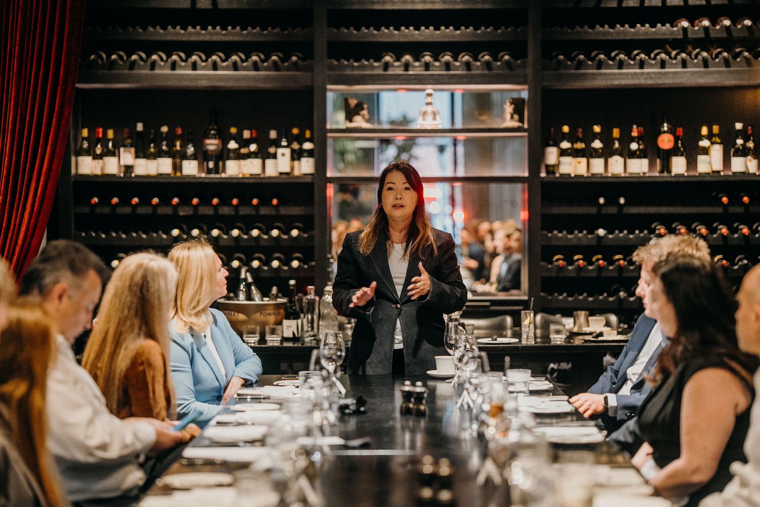 People gather around a long table for a wine tasting event in an elegant room with dark wood wine racks.