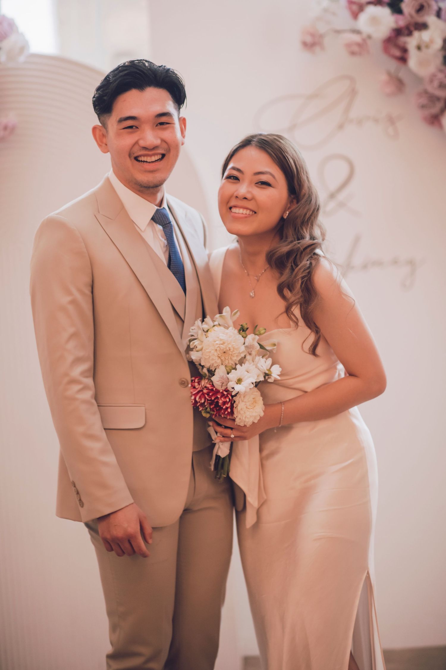 A couple in formal attire poses together at a wedding with beige and pink decor.