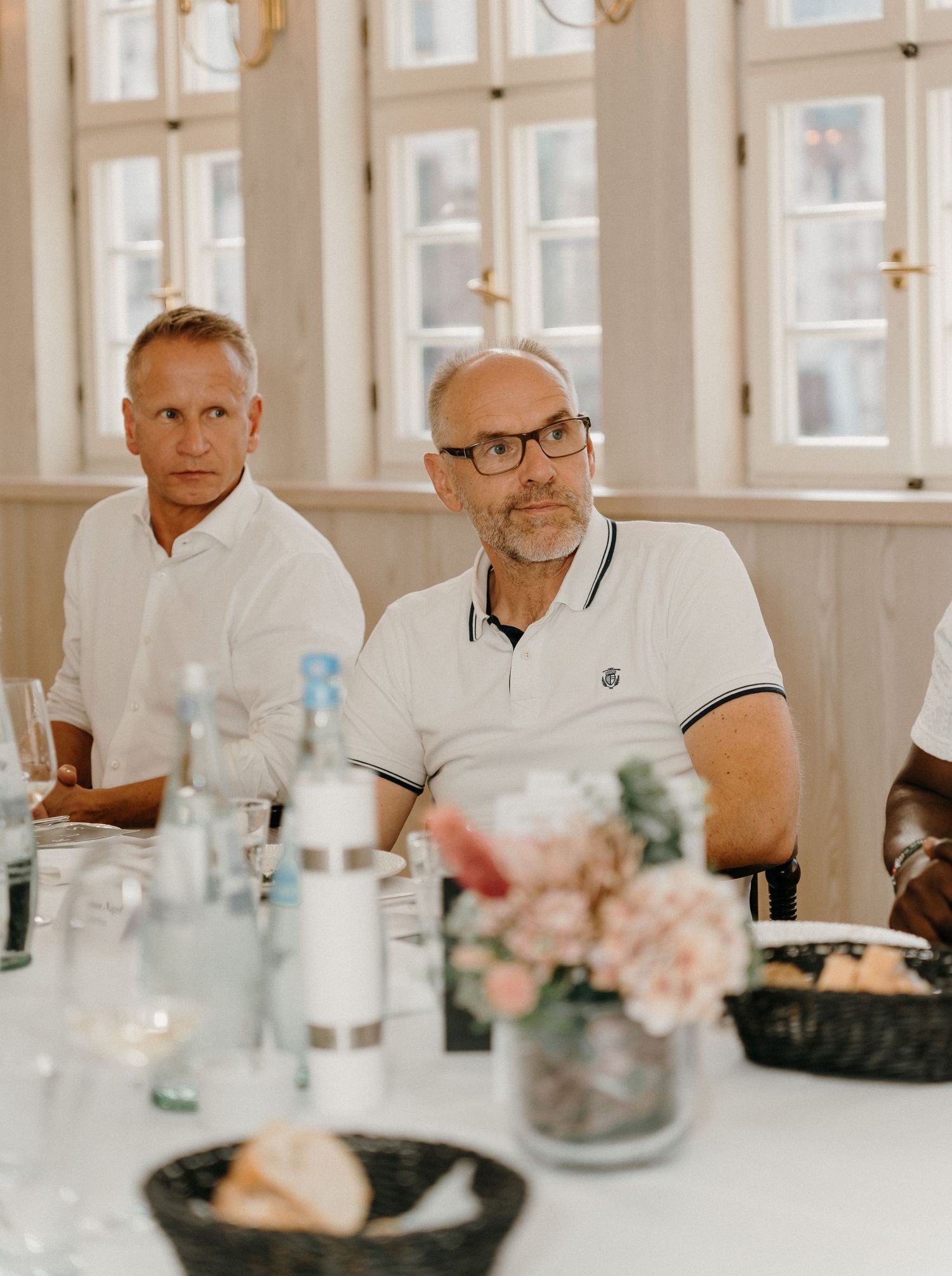 Two men in white shirts sit at a dining table with water bottles and flowers during a formal event.