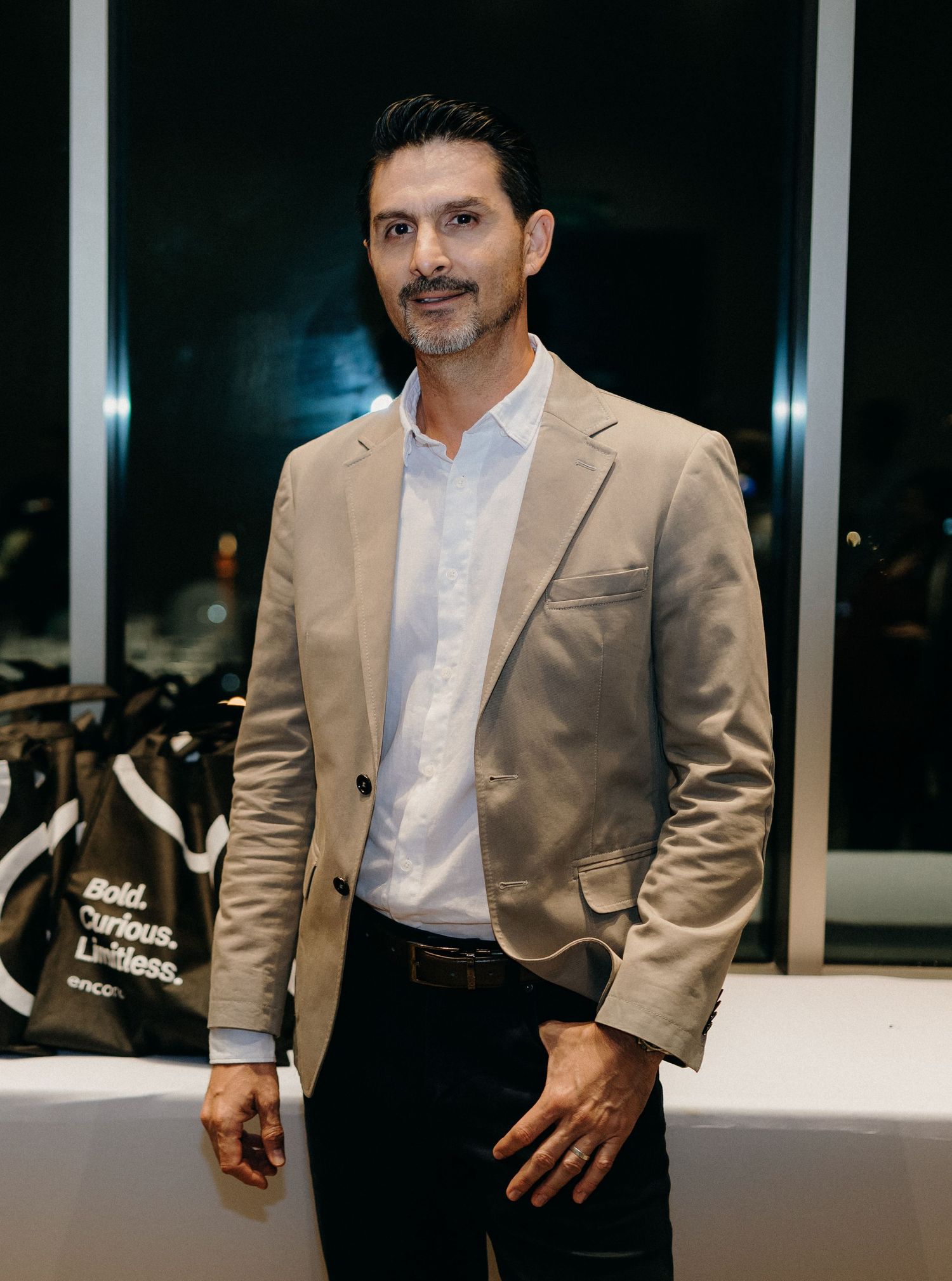 Professional wearing a tan suit jacket and white dress shirt poses in a modern office setting at night.