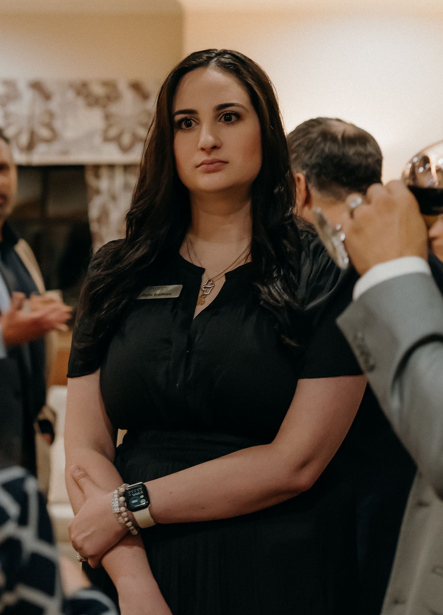 Professional in black attire stands confidently at an indoor event with others visible in background.