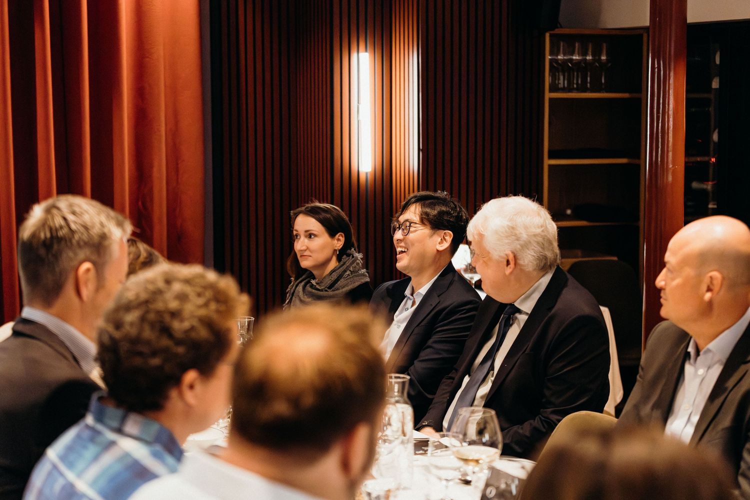 Business professionals seated around a long table during an evening meeting in an elegant wood-paneled conference room.