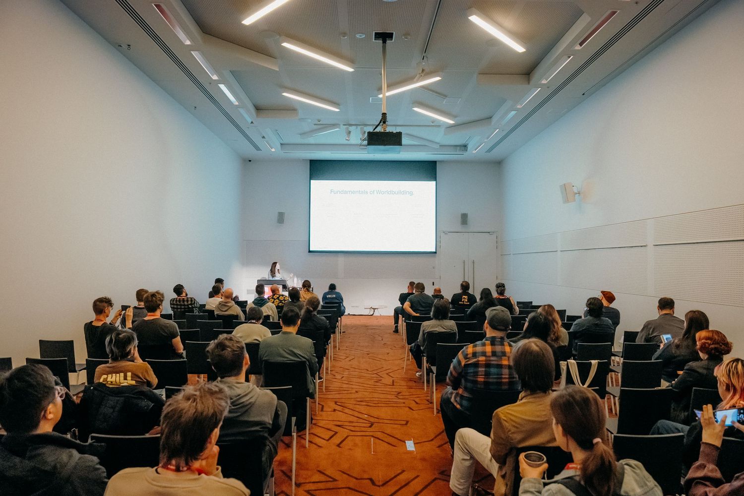 Students seated in rows in a modern classroom with white walls and fluorescent lighting during a lecture presentation.