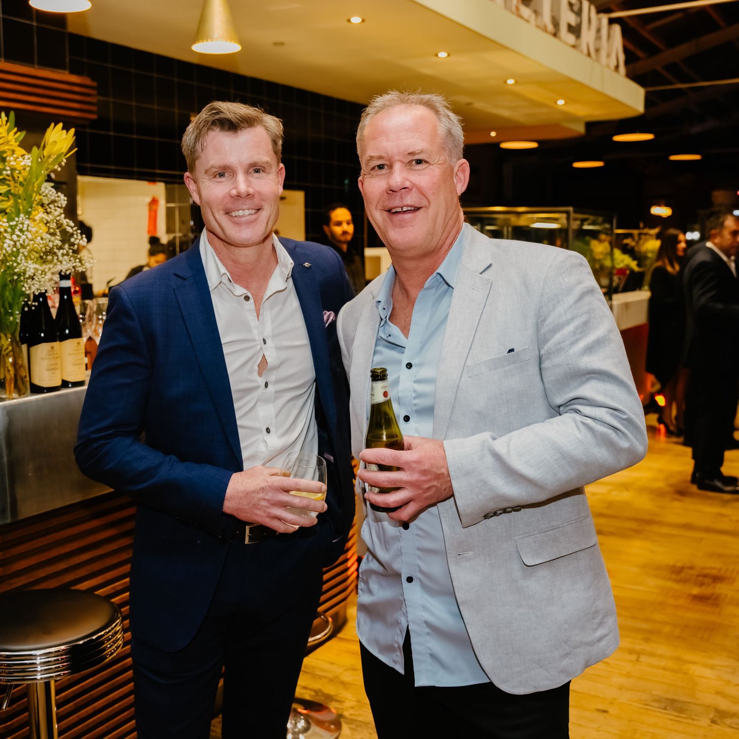 Two people in business attire share drinks and conversation at an indoor venue with warm lighting.