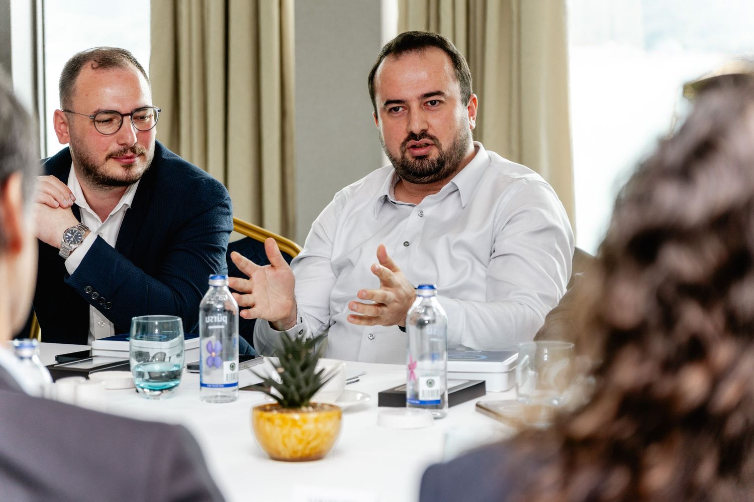 Two businessmen engaged in discussion at a conference table with water bottles and a pineapple centerpiece.