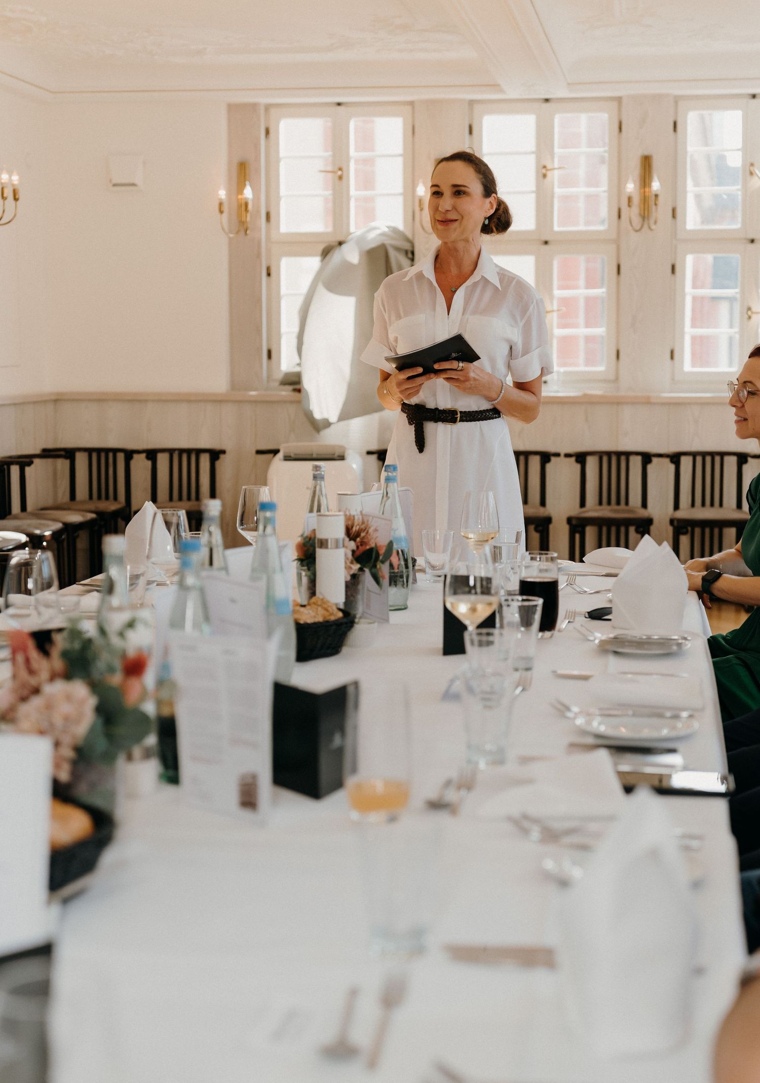 Elegant woman in white dress holding a book speaks at a formal dining event in a bright, decorated banquet hall.