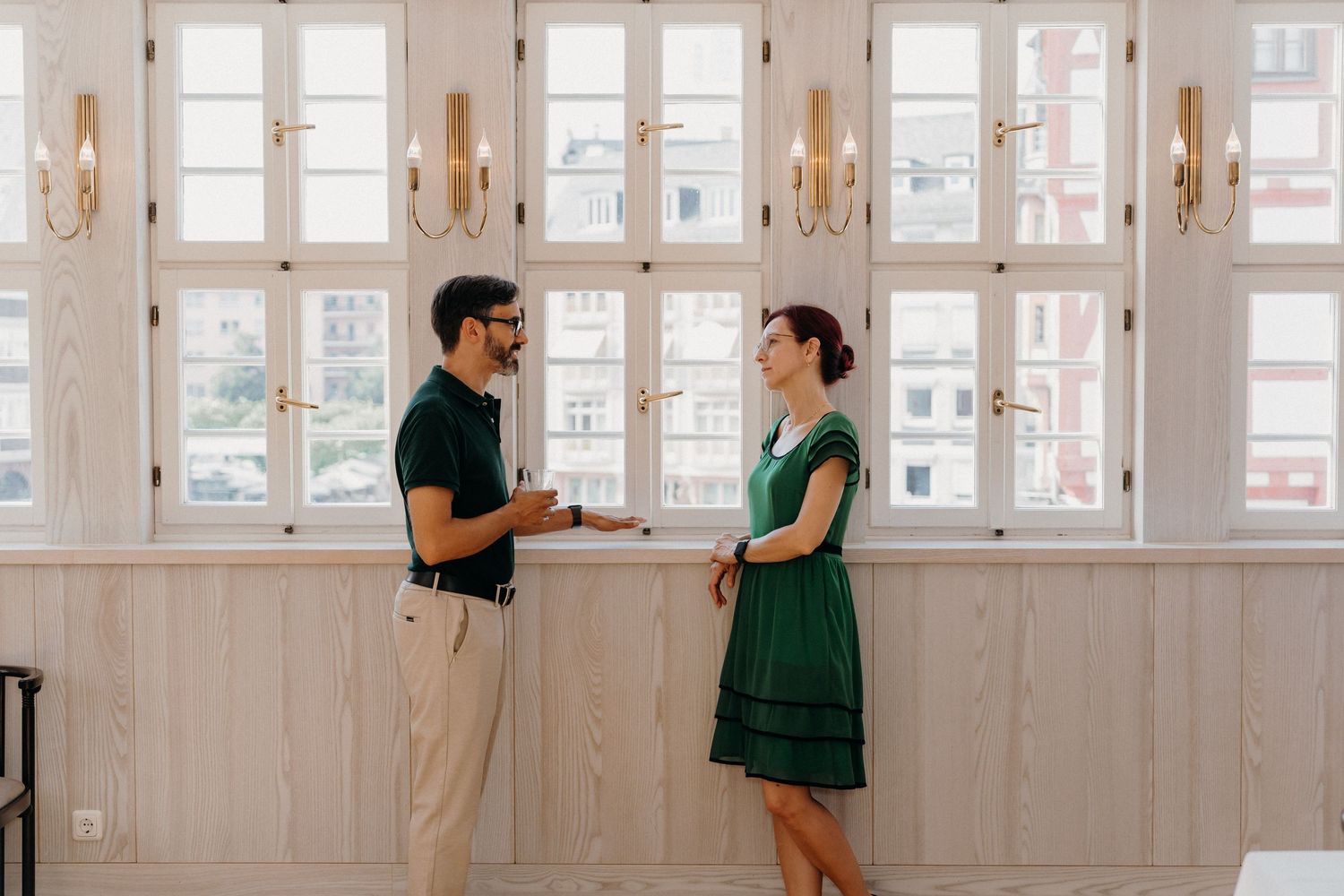 A man and woman in green outfits converse in a bright, elegant room with large windows and wall sconces.