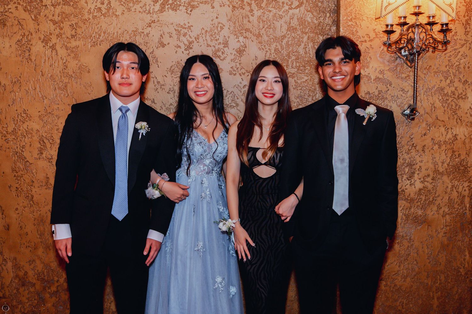 Four formally dressed people pose together at an elegant event with a warm-lit textured wall and chandelier behind them.