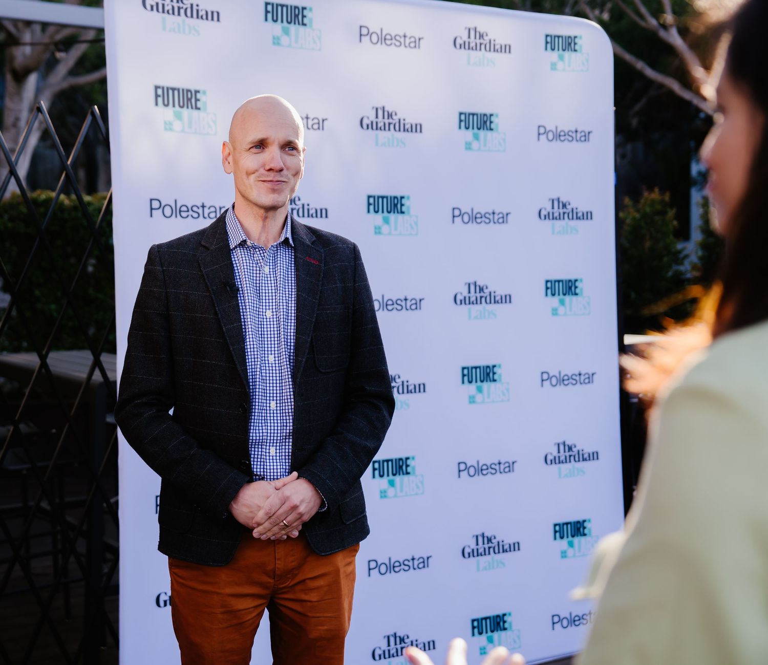 A person in a black blazer and striped shirt stands against a branded step-and-repeat backdrop at an event.