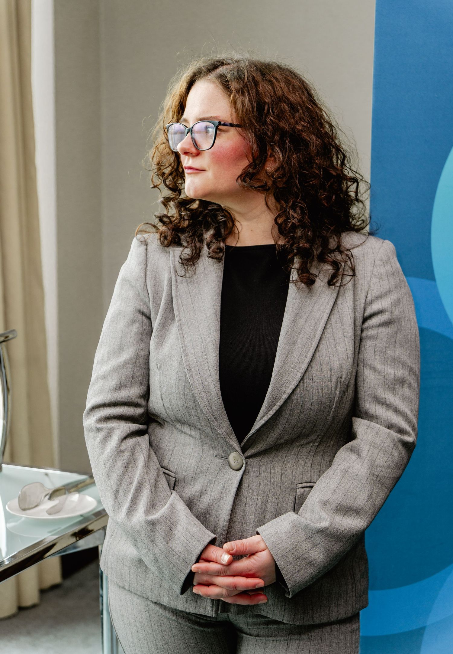 Professional woman in gray suit with curly hair and glasses standing confidently in a modern office setting.