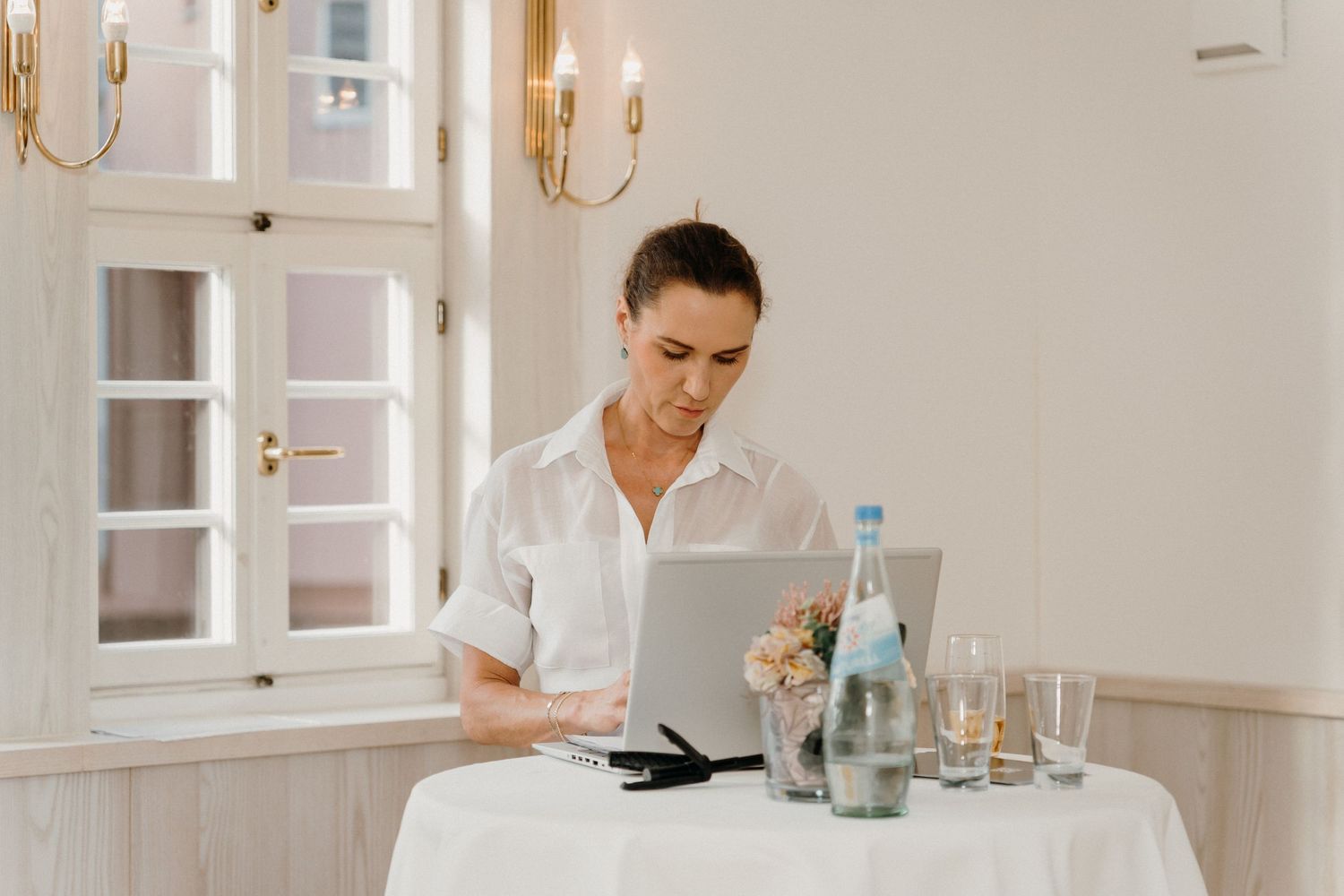 Professional woman in white shirt working on laptop at elegant table with water bottles and glasses.