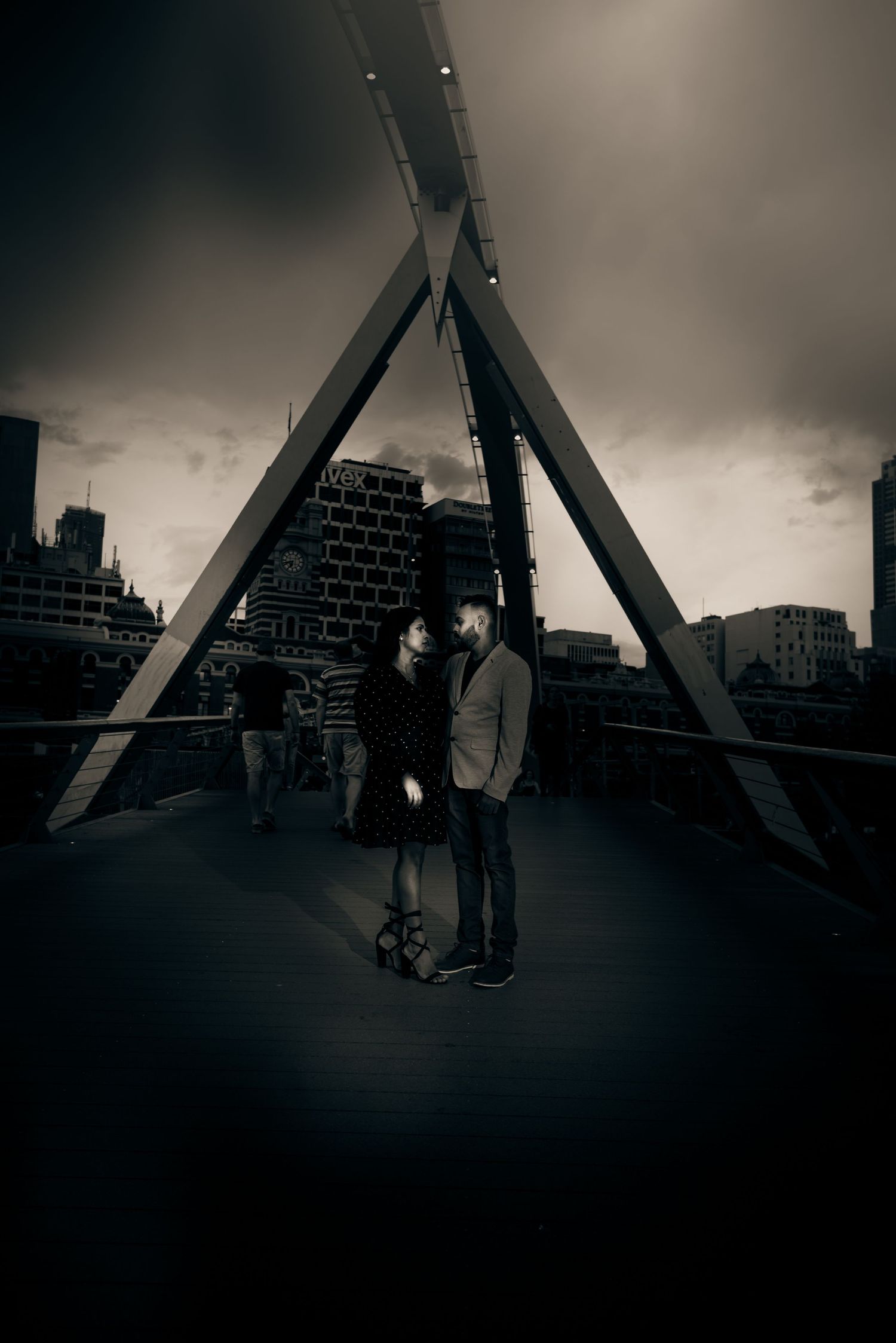 A moody black and white urban photograph shows two silhouettes on a pedestrian bridge with dramatic architectural beams.