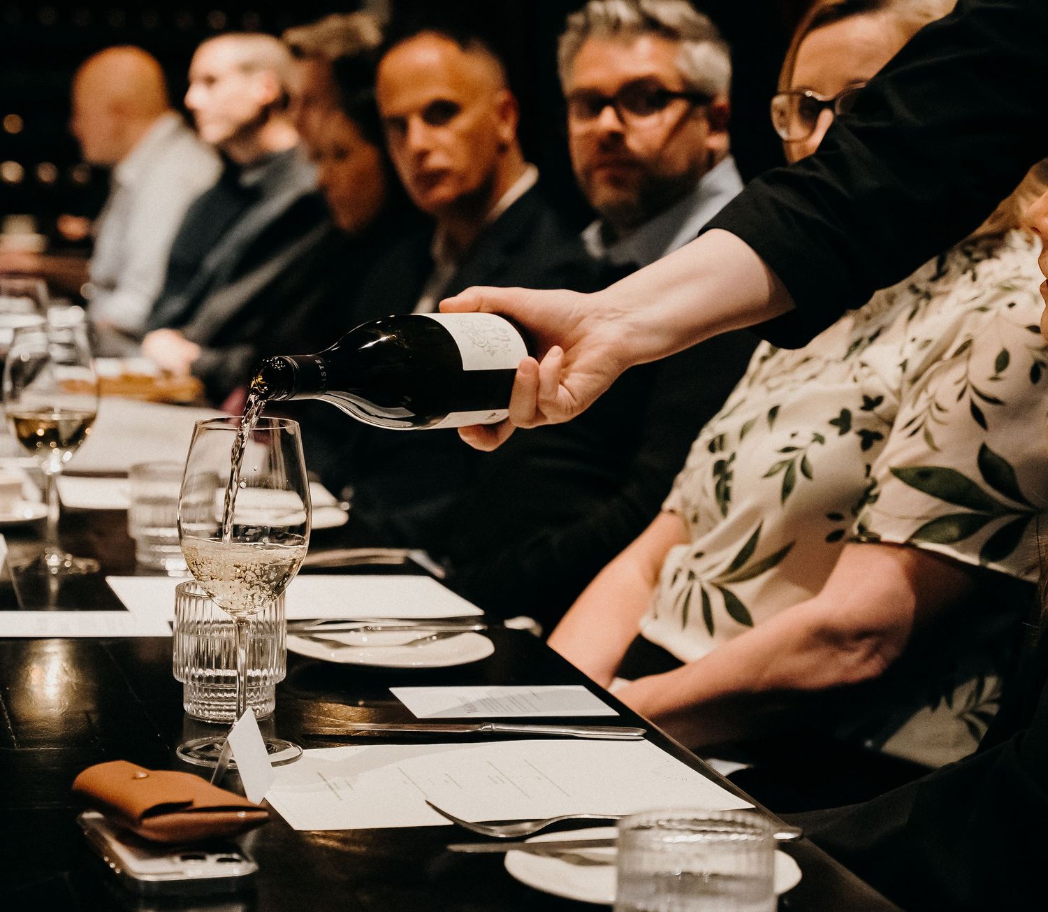 Hands serve elegant plated dishes at an upscale restaurant dinner service with glowing candlelight and white tablecloths.