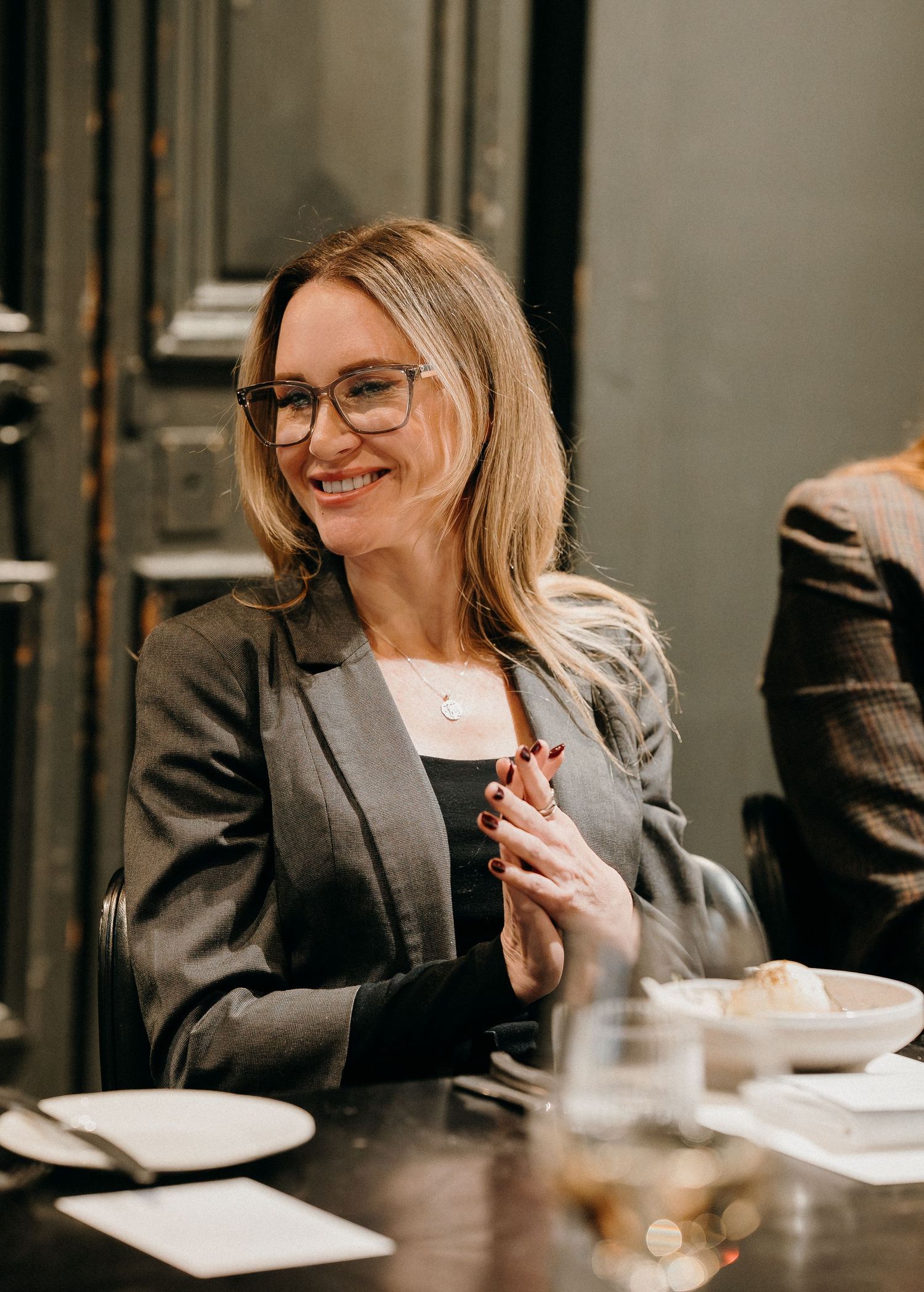 Professional in a gray blazer sits at a meeting table with paperwork and beverages while smiling warmly.