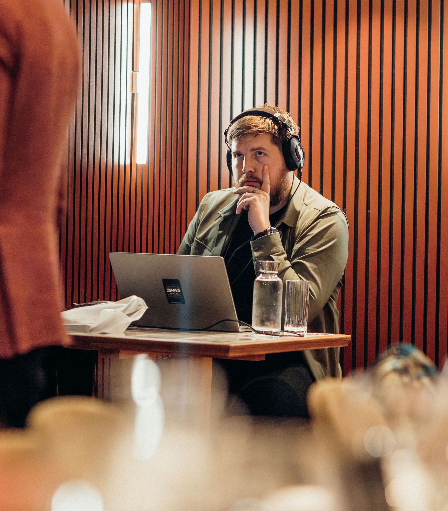 Someone wearing headphones works on a laptop at a desk in a wood-paneled room with a water bottle nearby.