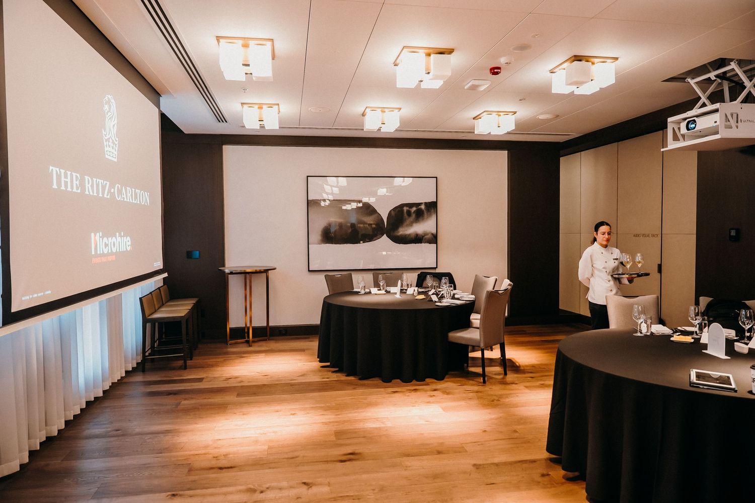 A modern conference room with wooden floors, black tables, and projector screens displaying business presentations.