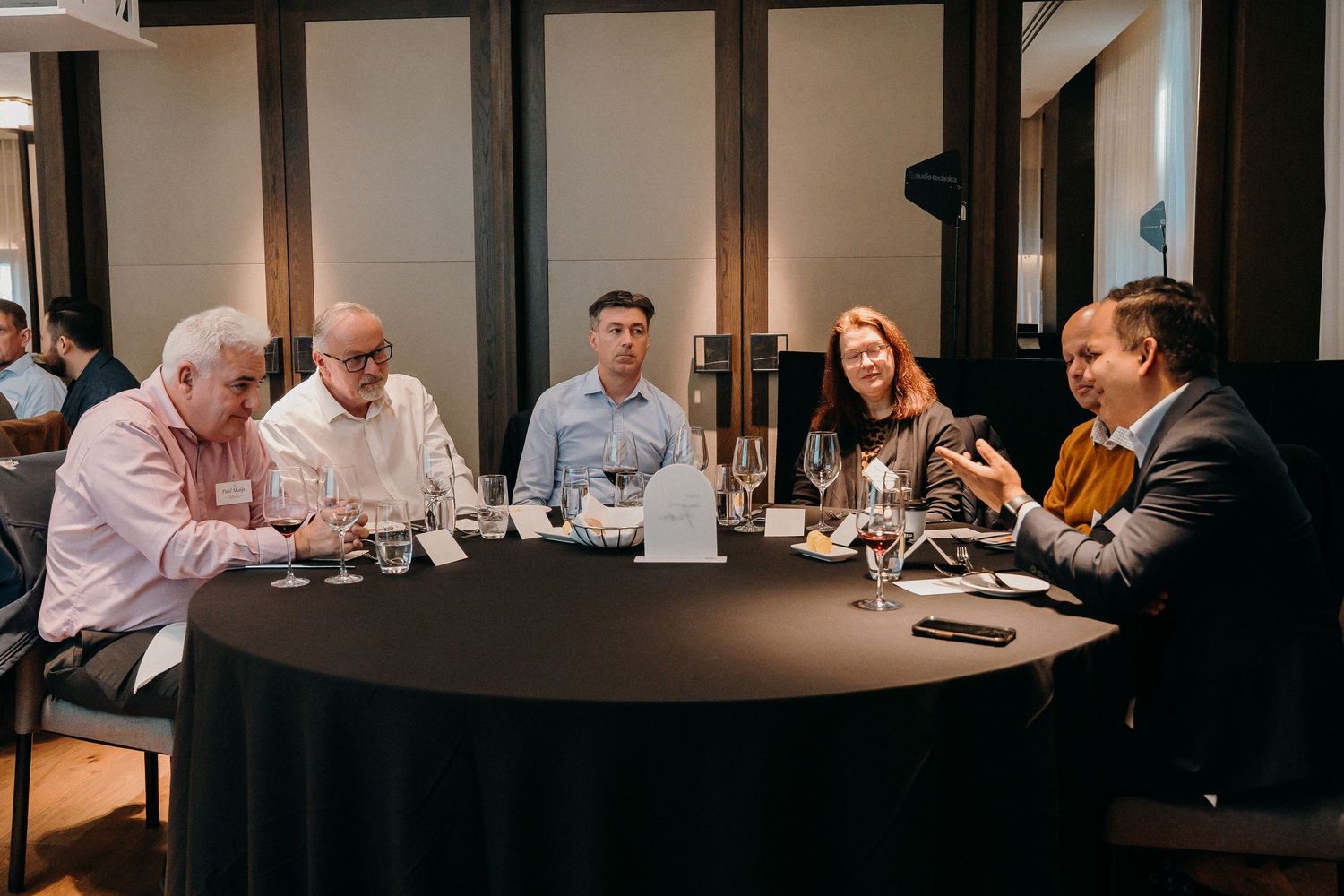 People seated around a dark round table with wine glasses having a business meeting or tasting event.