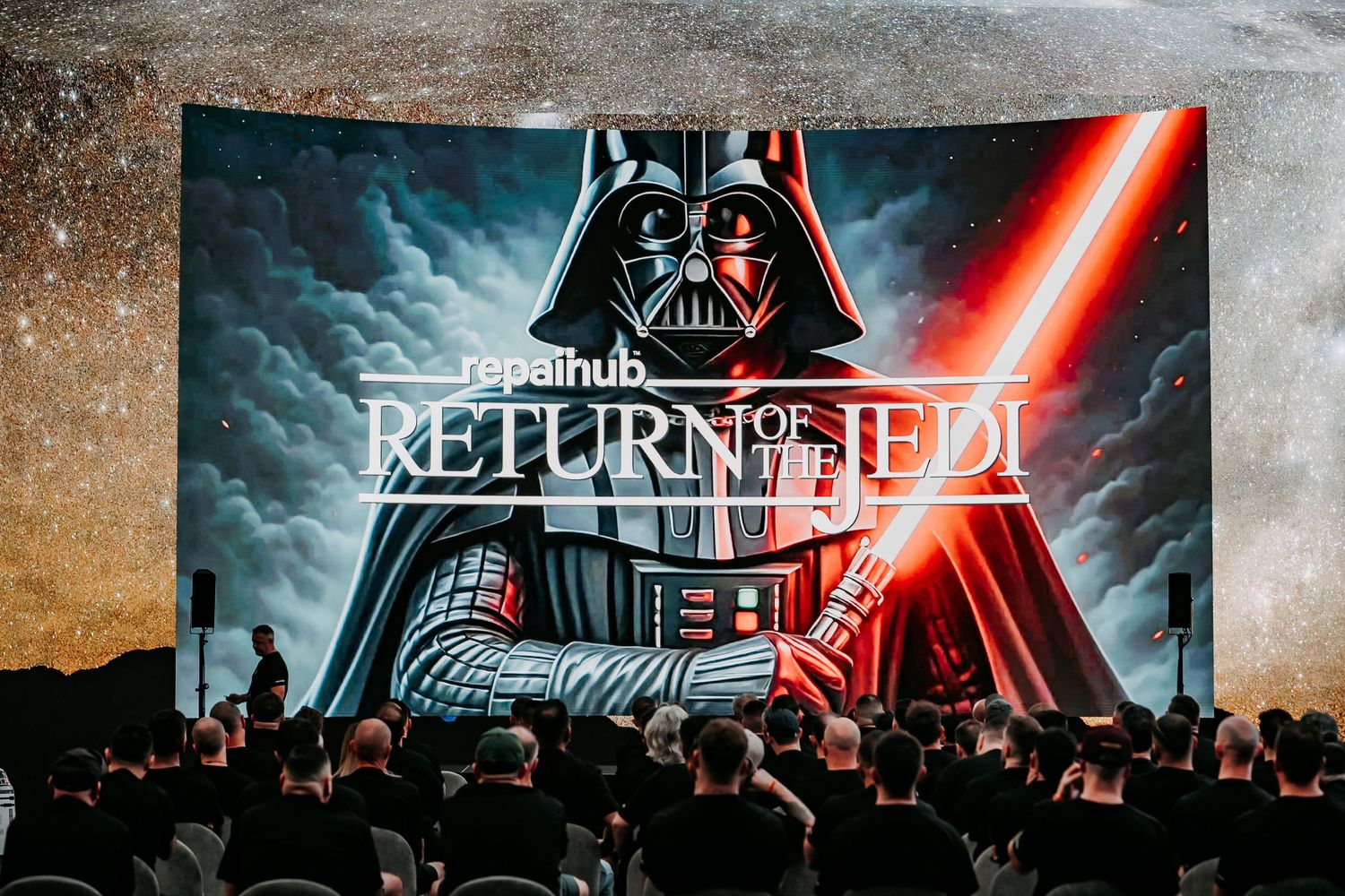 A movie theater audience watches Star Wars Return of the Jedi with Darth Vader and red lightsaber projected on screen.