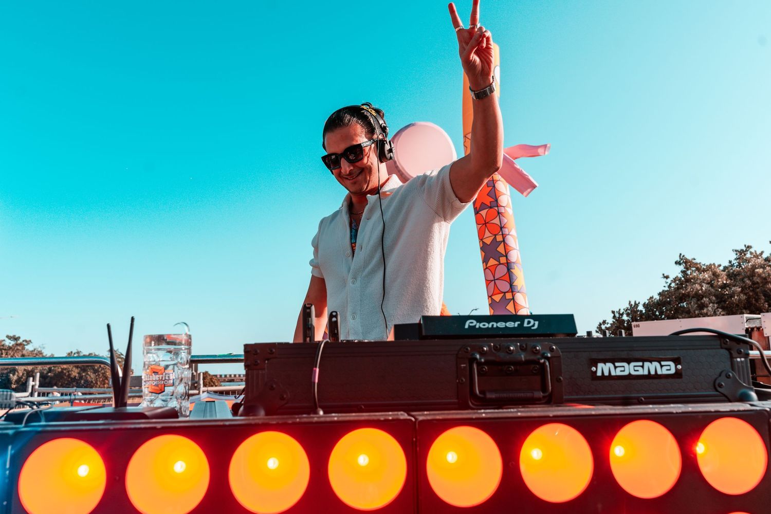 DJ performs at an outdoor event with orange glowing lights and turntables against a bright blue sky backdrop.