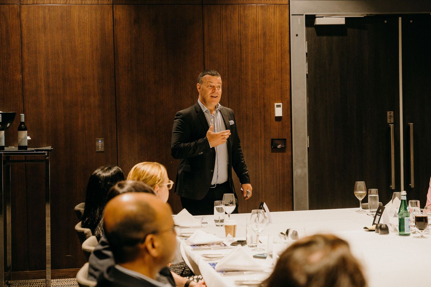 Business professional delivering a presentation to seated colleagues at a formal conference dining table in a modern boardroom.