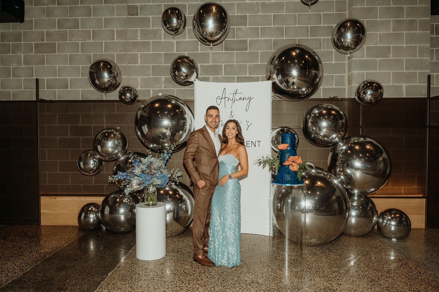 A couple stands in front of a modern balloon arch installation with silver balloons against a brick wall.