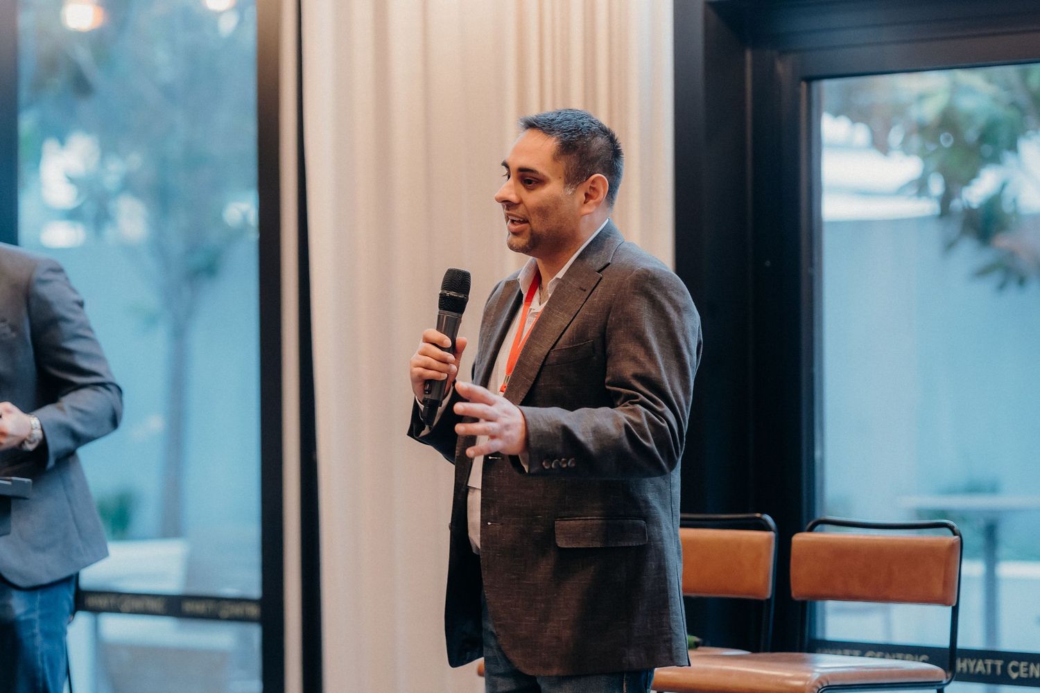 Professional in brown suit jacket speaks into microphone while gesturing during business presentation in office.