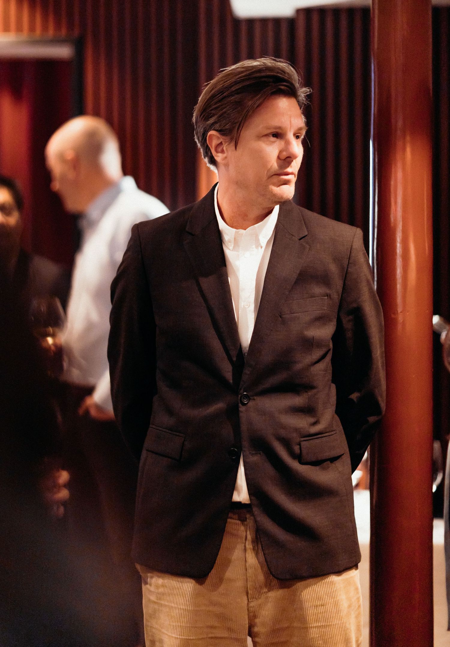 A person wearing a dark blazer and khaki pants stands in a dimly lit room with red walls and wooden pillars.