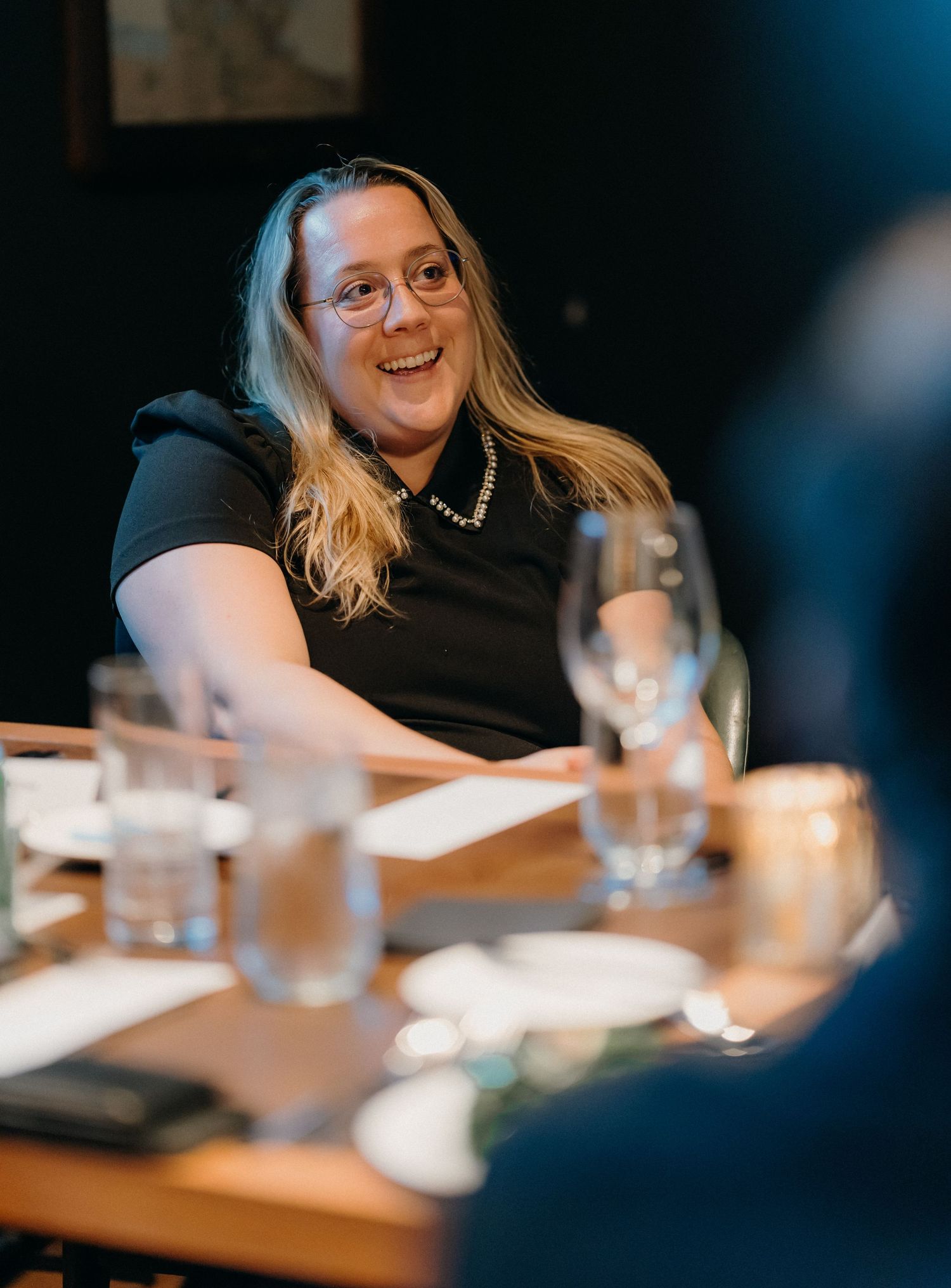 A person wearing glasses smiles and laughs during a meeting at a table with water glasses visible.