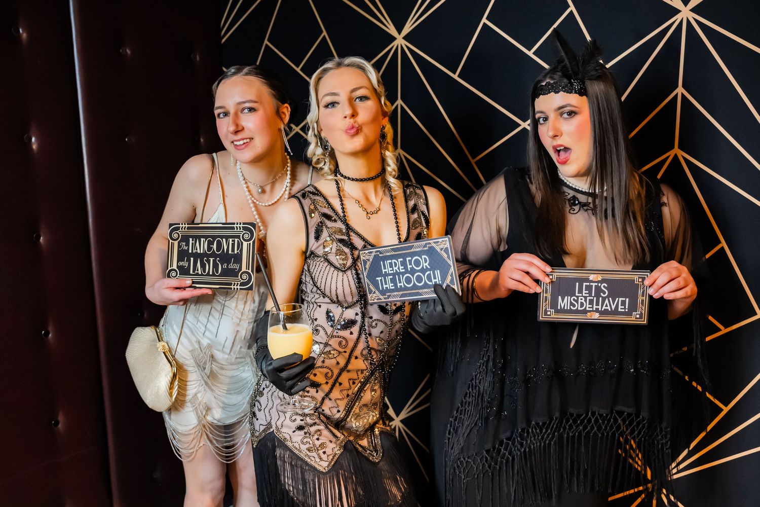 A group poses together with chalkboard signs at a party against a geometric gold backdrop.