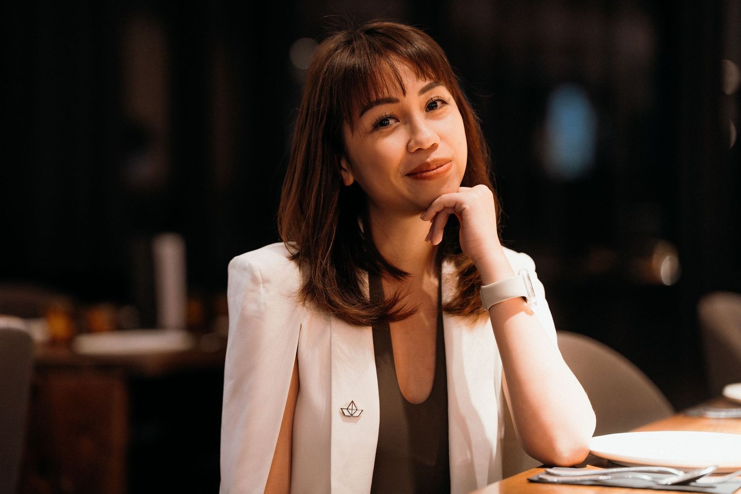 A person in a white blazer sits thoughtfully at a table in a dimly lit restaurant setting.