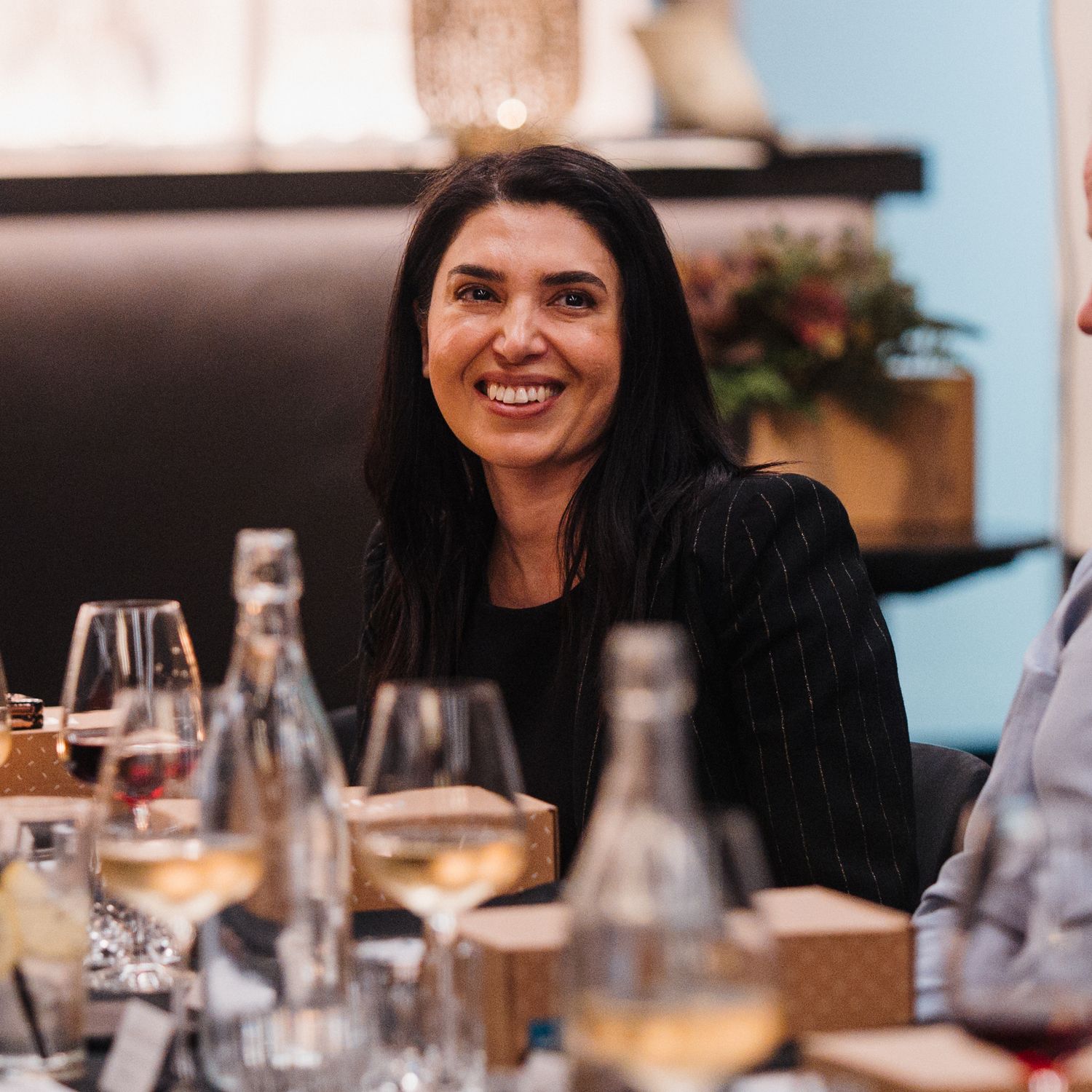 Someone with long dark hair smiles warmly while enjoying dinner with wine and water bottles on the table.