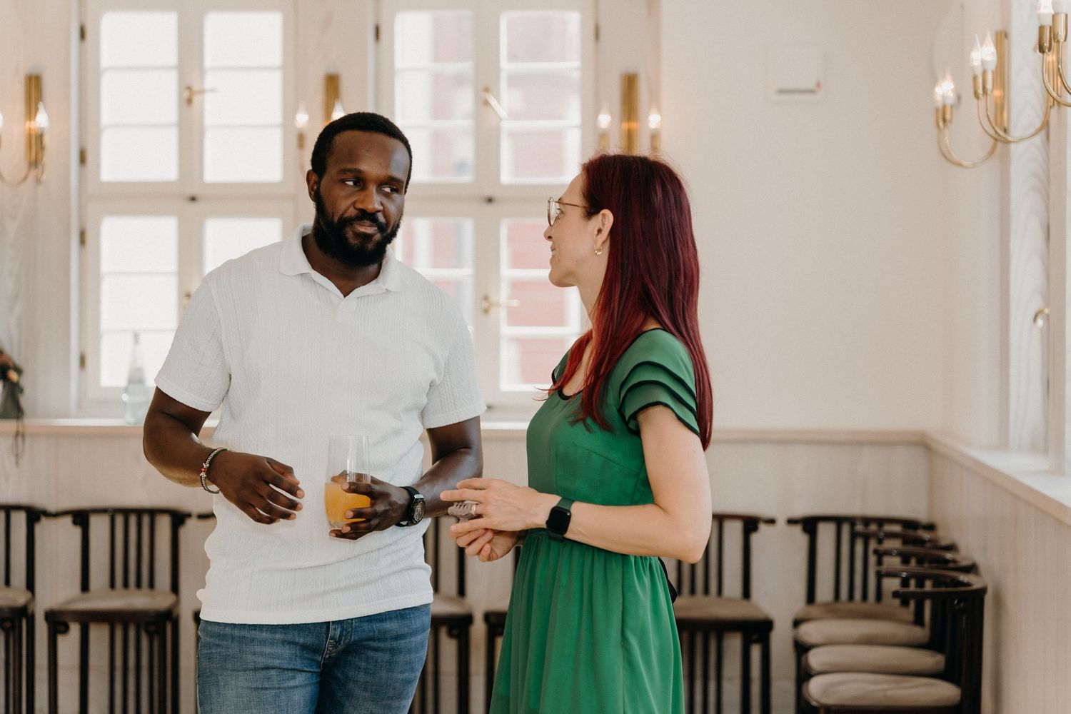 A Black man in a white shirt and a redhead woman in a green dress converse in an elegant dining room.
